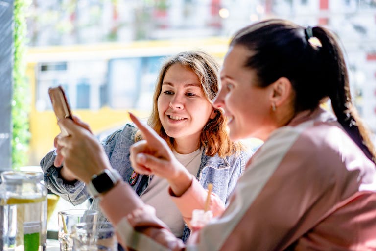 Smiling women in casual clothes using smartphone while sitting at table with glasses of water in cafe on summer day