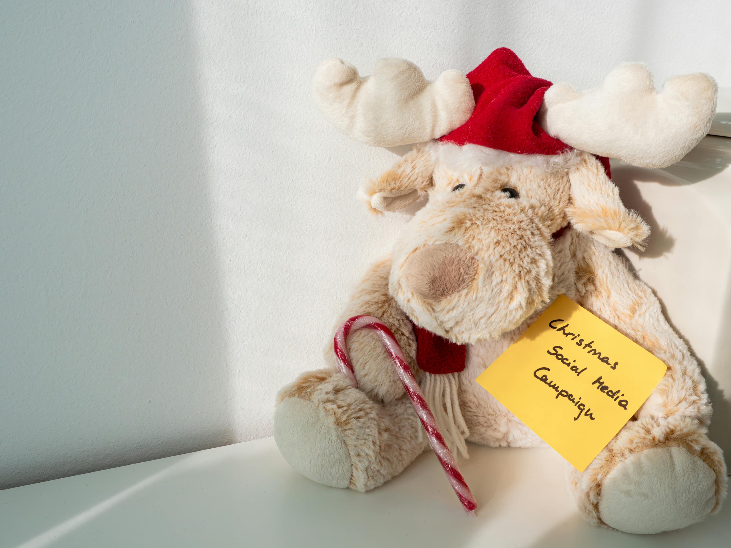 Plush reindeer toy wearing a Santa hat, holding a candy cane with a note reading 'Christmas Social Media Campaign'.
